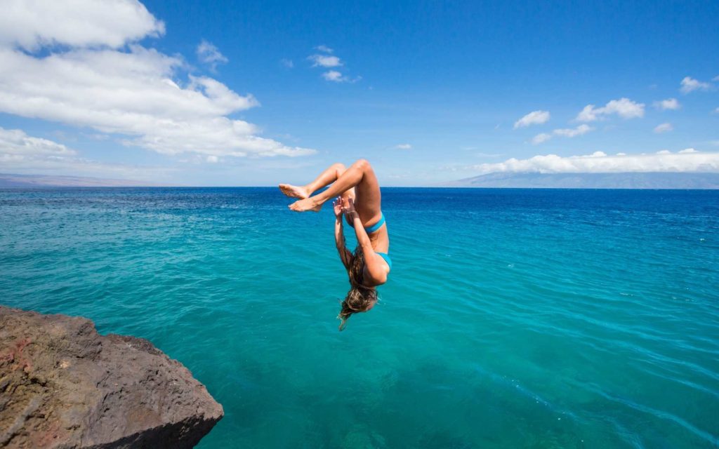 Image d'une femme courageuse qui ose sauter en backflip dans la mer avec le ciel bleu en fond et la mer turquoise